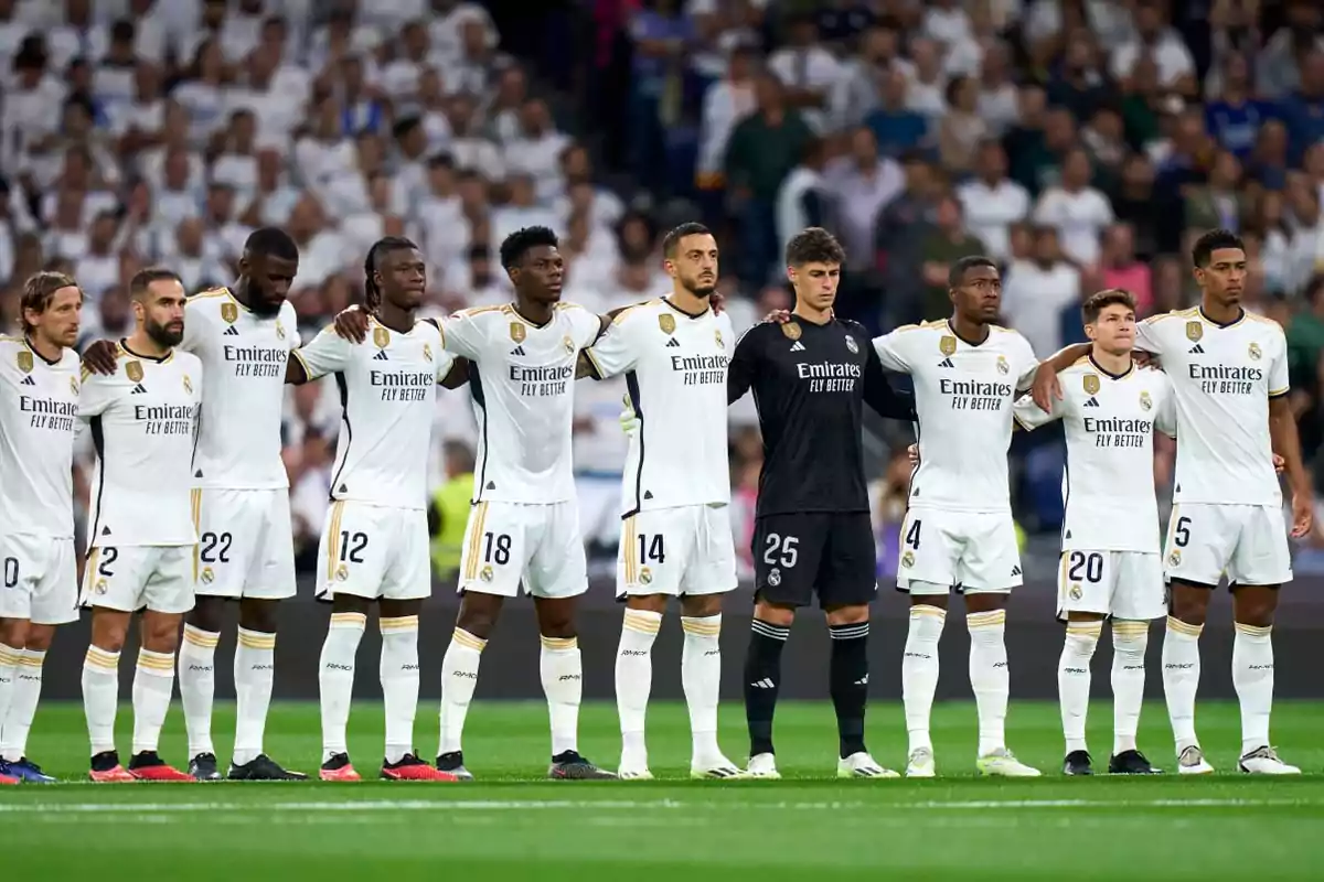 Jugadores de un equipo de fútbol alineados en el campo antes de un partido, todos con uniformes blancos excepto el portero que viste de negro, con los brazos sobre los hombros de sus compañeros.