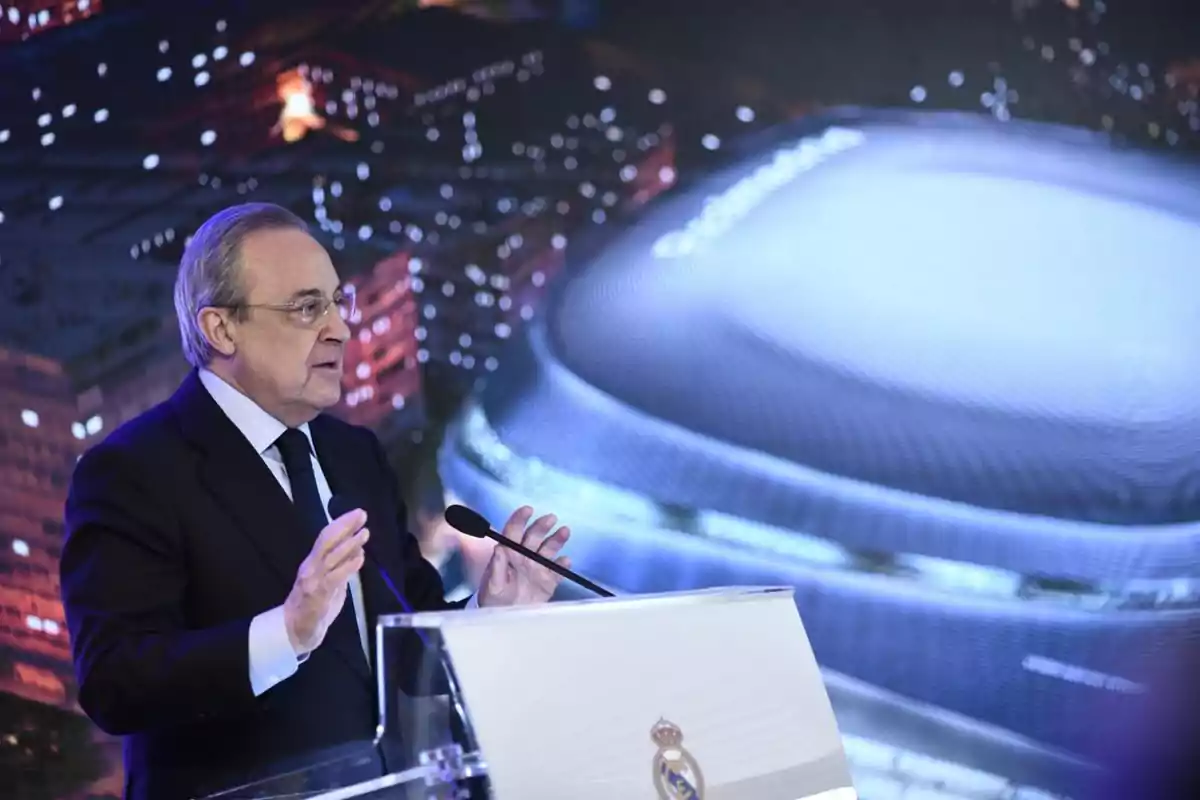 Un hombre de traje oscuro y corbata habla en un podio con el escudo del Real Madrid, con una imagen de un estadio moderno iluminado en el fondo.