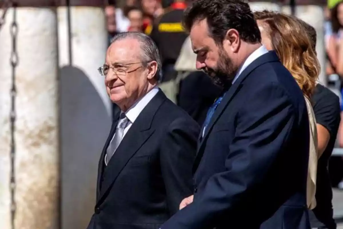 Dos hombres con trajes oscuros caminando juntos en un evento al aire libre.