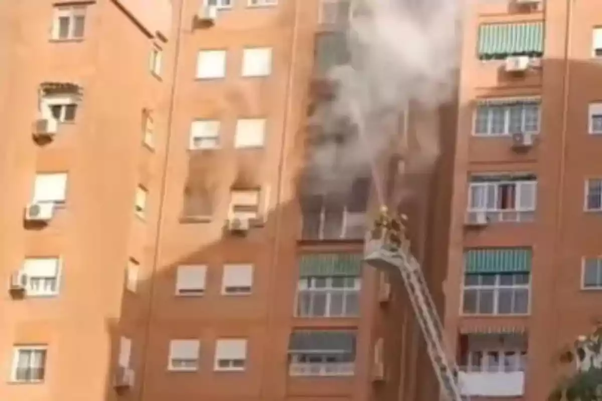 Un edificio de apartamentos de ladrillo con humo saliendo de una de las ventanas mientras los bomberos trabajan desde una escalera elevada. Un edificio de apartamentos de ladrillo con humo saliendo de una de las ventanas mientras los bomberos trabajan desde una escalera elevada.