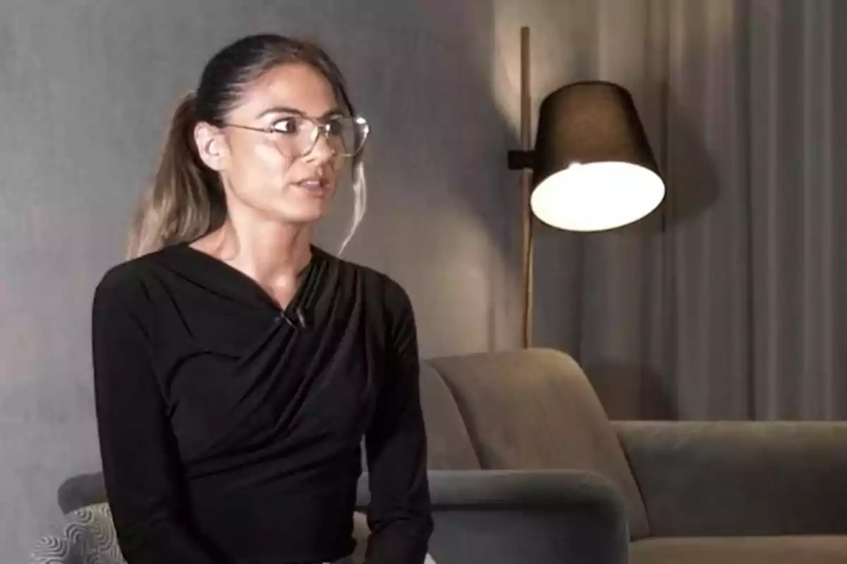 Una persona con gafas y cabello recogido está sentada en un sofá frente a una lámpara encendida en una habitación con paredes grises. Una persona con gafas y cabello recogido está sentada en un sofá frente a una lámpara encendida en una habitación con paredes grises.
