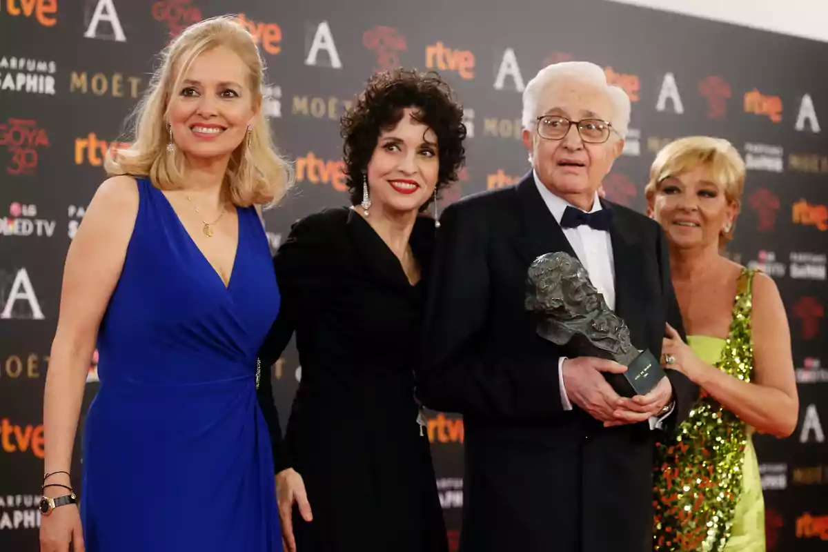 Un grupo de personas posando en una alfombra roja, una de ellas sostiene un trofeo. Un grupo de personas posando en una alfombra roja, una de ellas sostiene un trofeo.