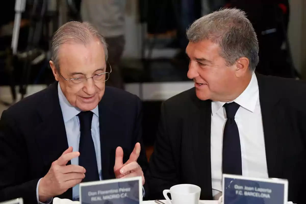 Dos hombres con trajes oscuros conversan en una mesa, uno de ellos tiene una placa que dice "Don Florentino Pérez REAL MADRID C.F." y el otro tiene una placa que dice "Don Joan Laporta F.C. BARCELONA".