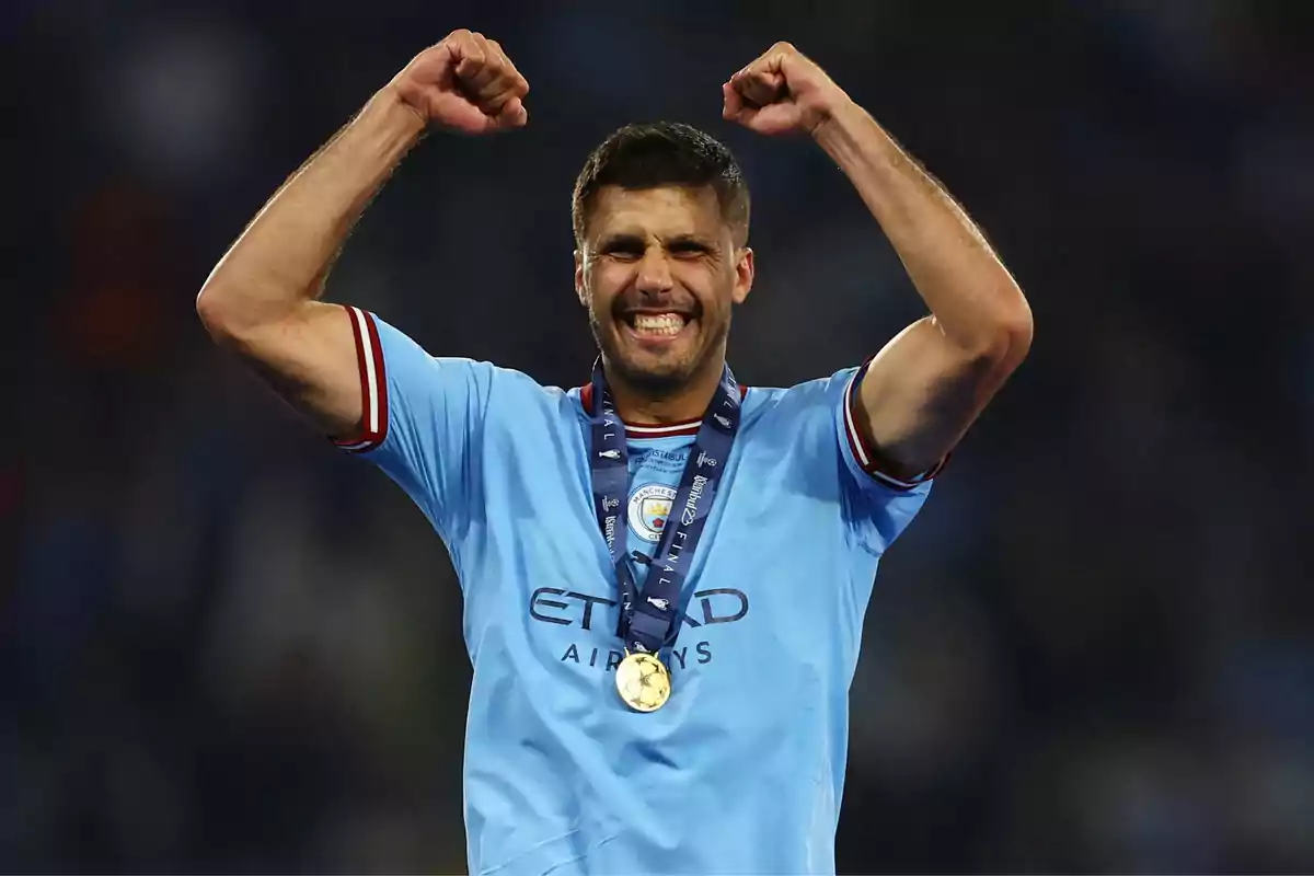 Jugador de fútbol celebrando con los brazos levantados y una medalla de oro alrededor del cuello, vistiendo una camiseta azul claro del Manchester City.