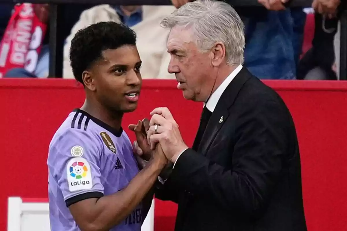 Un jugador de fútbol con uniforme morado interactúa con un hombre mayor vestido de traje negro en un estadio.