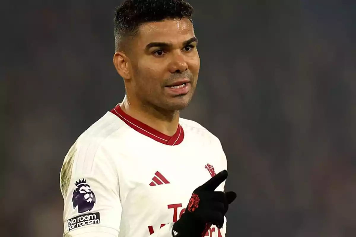 Un jugador de fútbol con uniforme blanco y detalles rojos, guantes negros y un parche de la Premier League en la manga, gesticulando durante un partido.