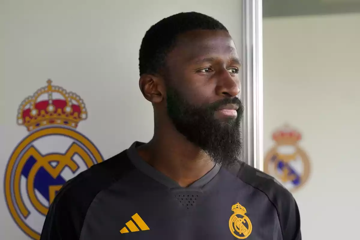 Un jugador de fútbol con la camiseta de entrenamiento del Real Madrid frente a un fondo con el escudo del equipo.