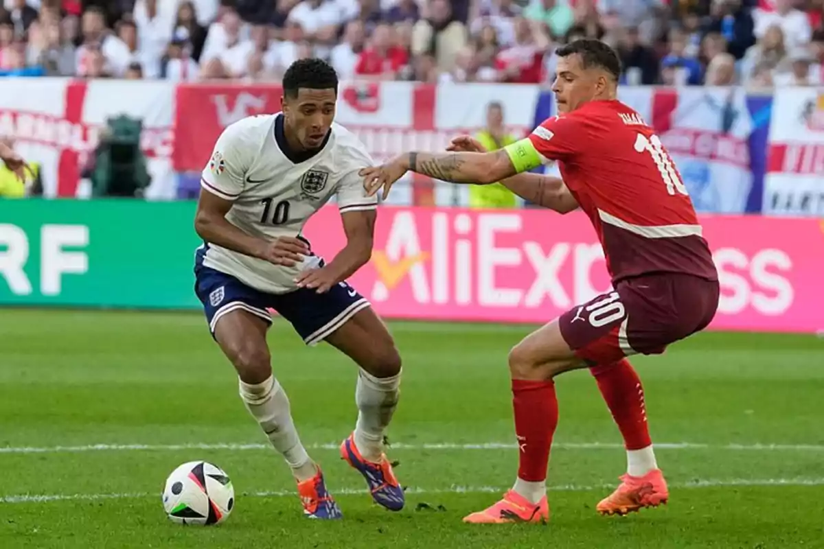 Dos jugadores de fútbol compiten por el balón en un partido, uno con uniforme blanco y el otro con uniforme rojo, mientras el público observa desde las gradas.