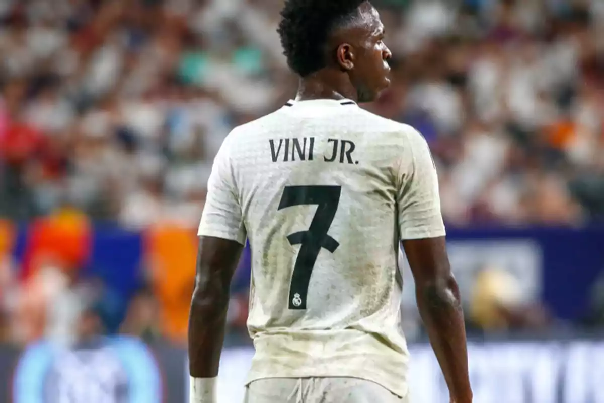 Jugador de fútbol con camiseta blanca y el nombre "VINI JR." y el número 7 en la espalda, en un estadio lleno de espectadores.