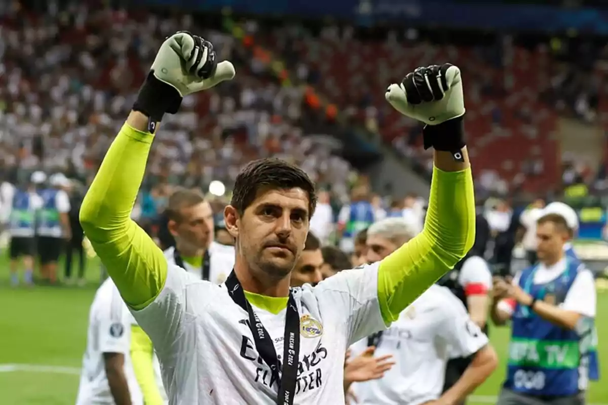 Un jugador de fútbol con uniforme blanco y guantes de portero levanta los brazos en señal de celebración en un estadio lleno de espectadores.