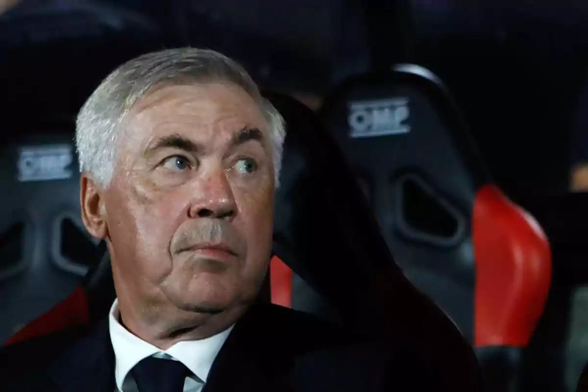 Hombre de cabello canoso sentado en un asiento deportivo, mirando hacia un lado.