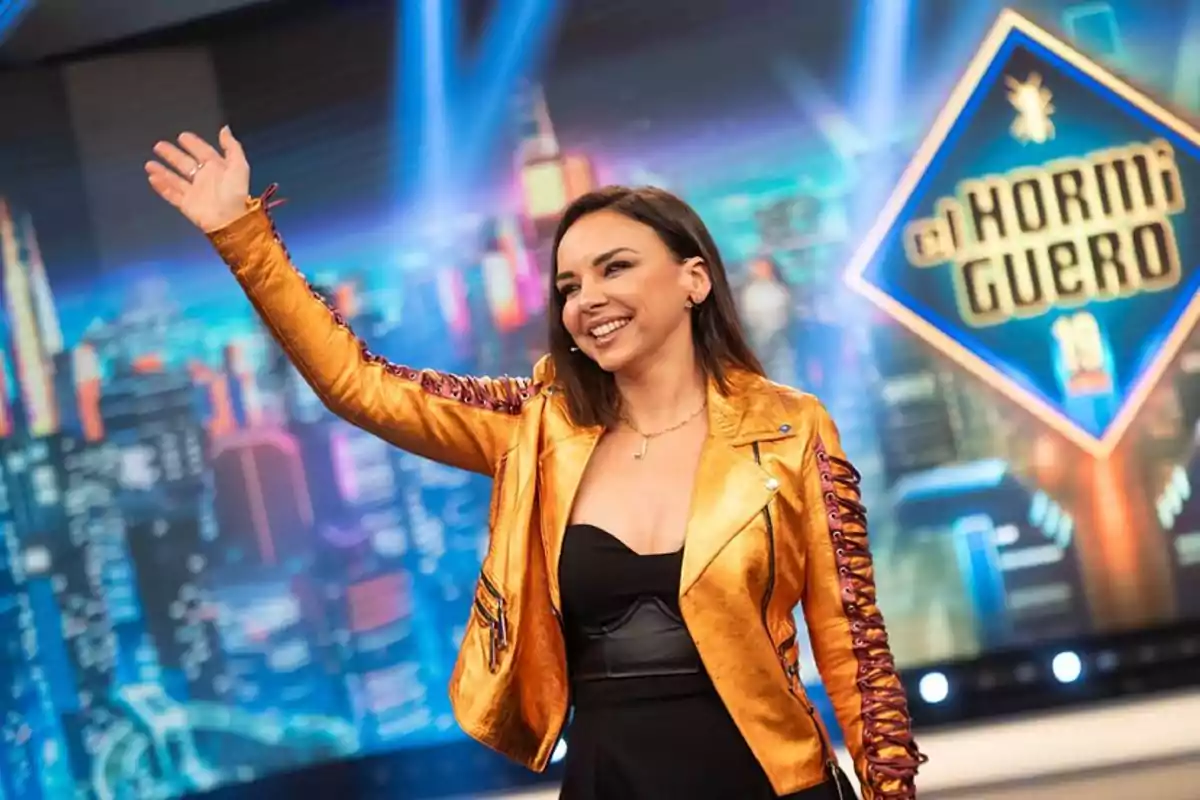 Una persona sonriente con chaqueta dorada levanta la mano en un set de televisión colorido.