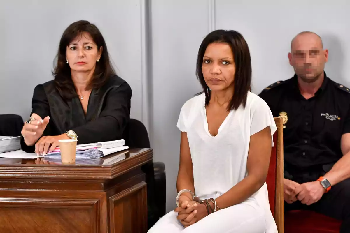 Una mujer con camiseta blanca está sentada junto a una mesa en un entorno formal, acompañada por una persona con uniforme de policía y otra persona con vestimenta profesional. Una mujer con camiseta blanca está sentada junto a una mesa en un entorno formal, acompañada por una persona con uniforme de policía y otra persona con vestimenta profesional.