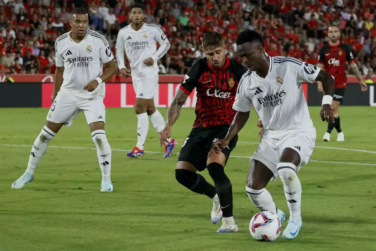 Jugadores de fútbol de dos equipos diferentes compiten por el balón en un partido, uno de los equipos viste de blanco y el otro de negro con rojo, mientras el público observa desde las gradas.