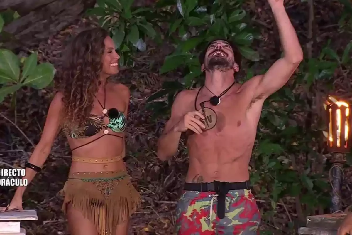 Dos personas sonríen en un entorno selvático, una mujer lleva un bikini y una falda con flecos, mientras que el hombre está sin camisa y levanta el brazo. Dos personas sonríen en un entorno selvático, una mujer lleva un bikini y una falda con flecos, mientras que el hombre está sin camisa y levanta el brazo.