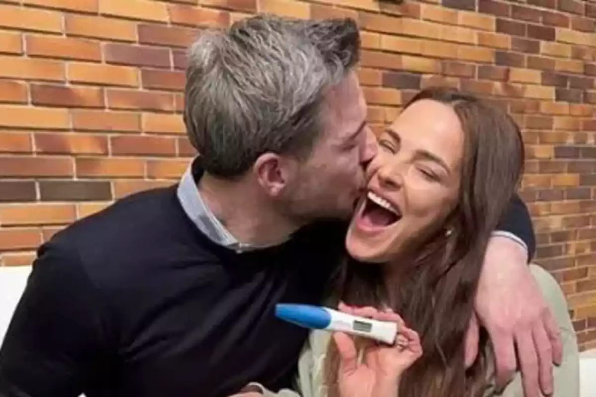 Una pareja sonriente se abraza mientras la mujer sostiene una prueba de embarazo positiva.