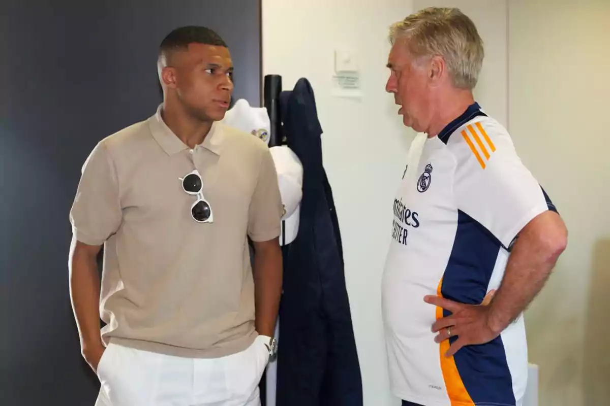 Dos hombres conversando, uno con camiseta beige y gafas de sol colgadas y el otro con camiseta deportiva blanca.