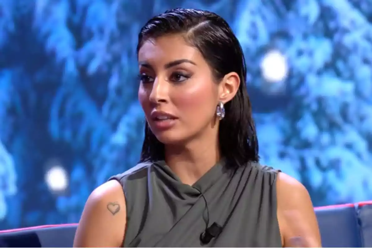 Mujer con cabello oscuro y mojado, usando un vestido gris y aretes brillantes, sentada frente a un fondo azul. Mujer con cabello oscuro y mojado, usando un vestido gris y aretes brillantes, sentada frente a un fondo azul.