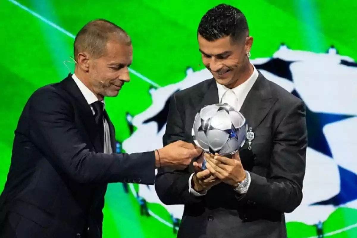 Dos hombres en traje sosteniendo un trofeo de fútbol en un escenario con fondo verde.