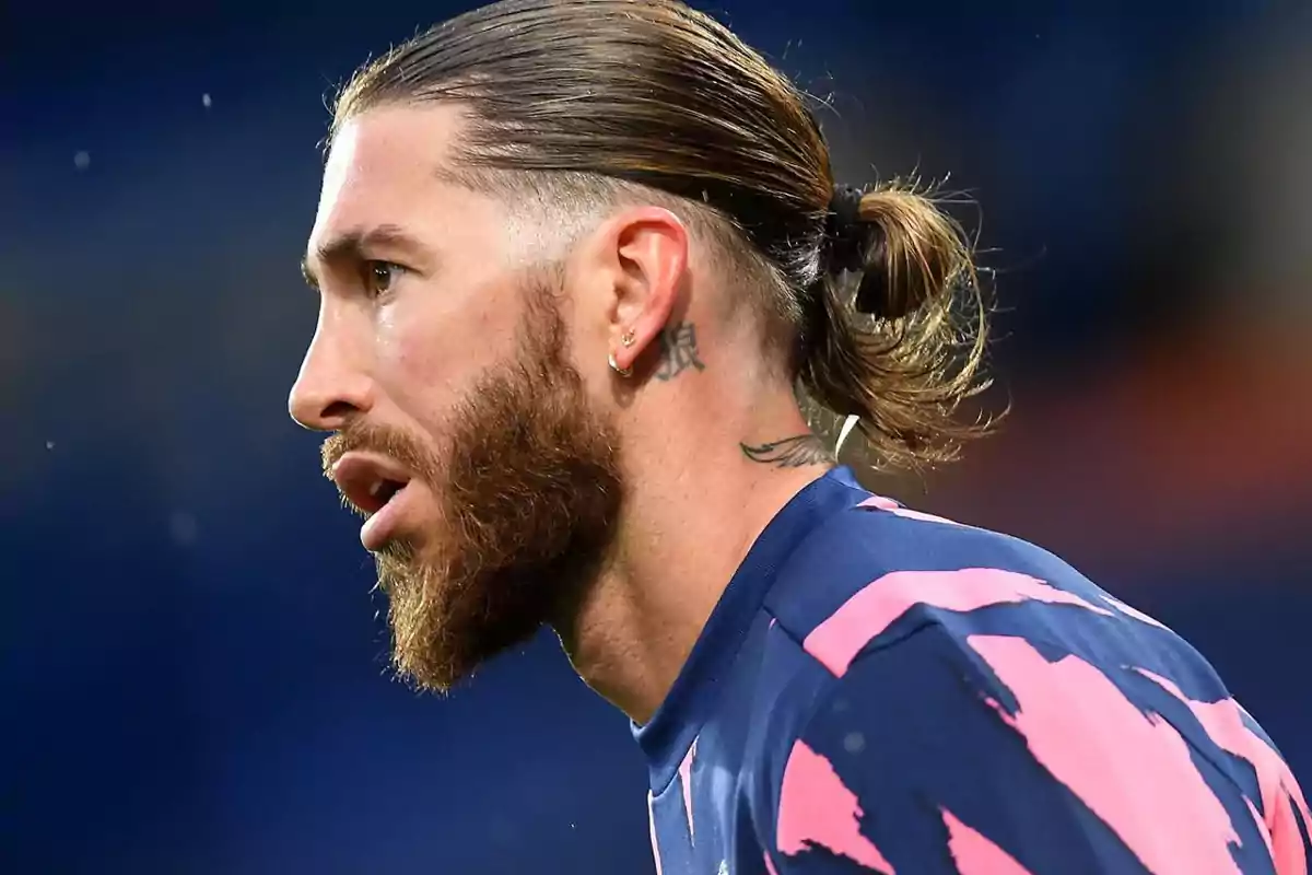 Un hombre con barba y cabello recogido en una cola de caballo, usando una camiseta deportiva azul con detalles rosados.