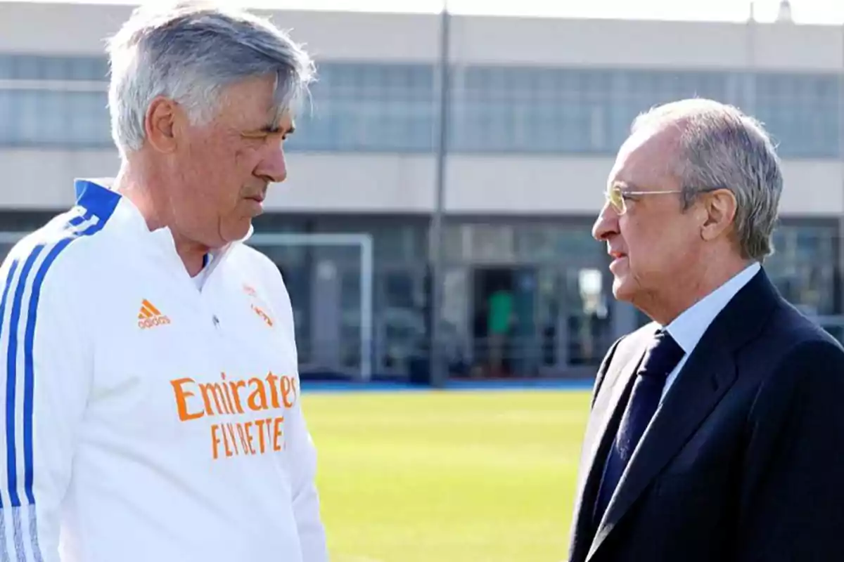 Dos hombres conversando en un campo de entrenamiento de fútbol.