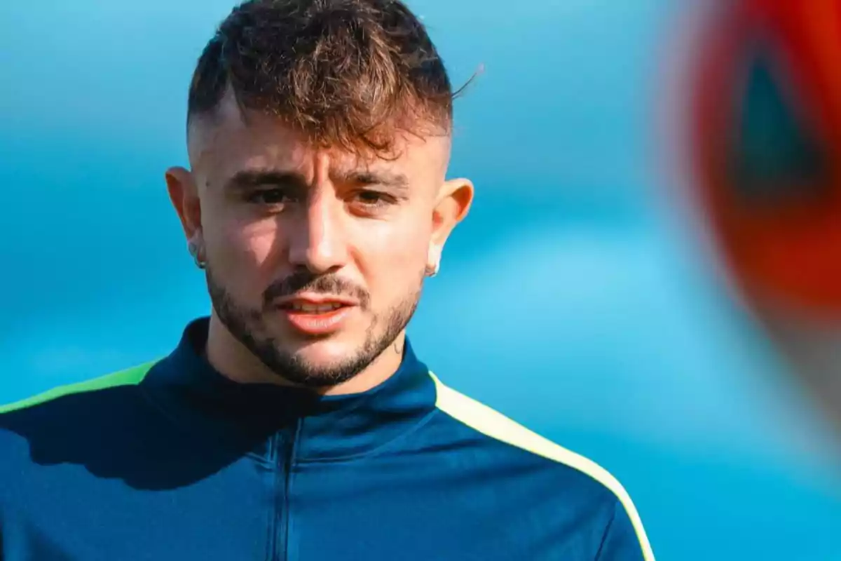 Un hombre con barba y cabello corto, vestido con una chaqueta deportiva azul con detalles en verde, mirando hacia adelante con un fondo azul difuminado.