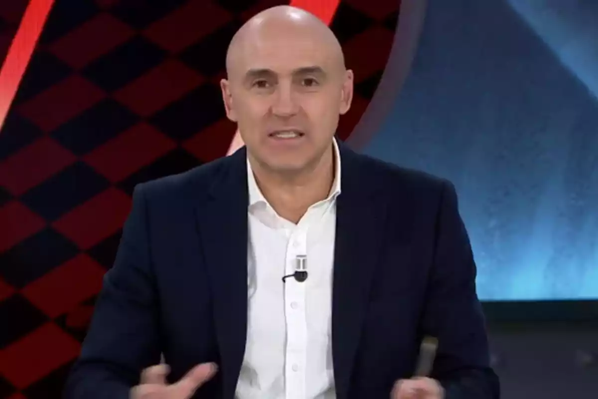 Un hombre calvo con traje oscuro y camisa blanca hablando en un programa de televisión con un fondo de patrón geométrico rojo y negro.