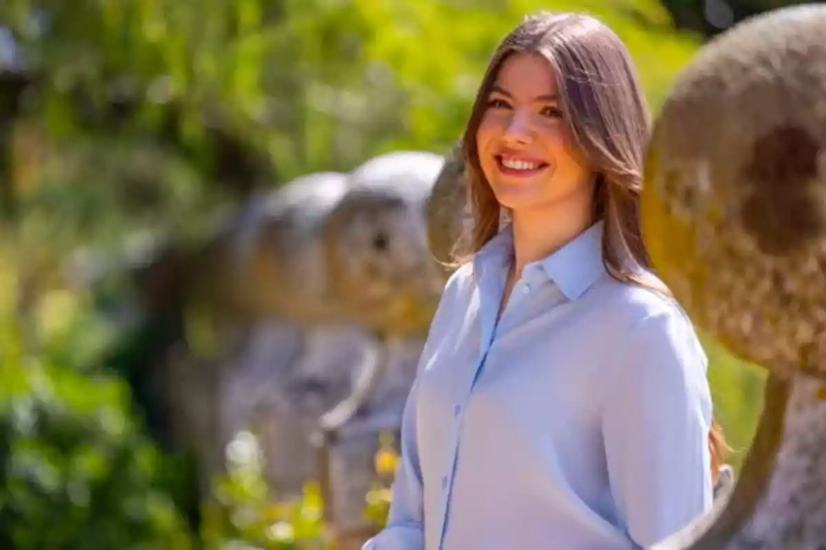 Una mujer sonriente con cabello largo y castaño lleva una camisa azul claro mientras está de pie en un entorno al aire libre con vegetación y esculturas de piedra en el fondo. Una mujer sonriente con cabello largo y castaño lleva una camisa azul claro mientras está de pie en un entorno al aire libre con vegetación y esculturas de piedra en el fondo.