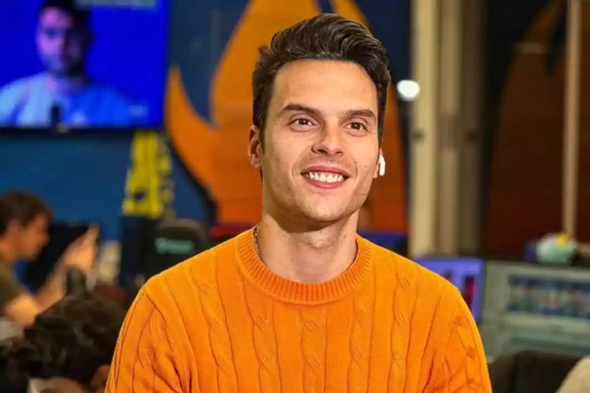 Persona sonriendo con un suéter naranja en un entorno de oficina con pantallas y personas de fondo.