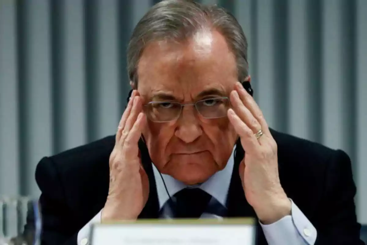 Hombre con gafas y traje oscuro, tocándose la cabeza con ambas manos, en una conferencia.