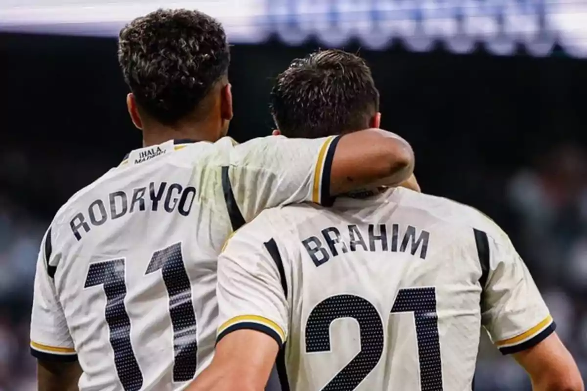 Dos jugadores de fútbol con camisetas blancas, uno con el número 11 y el nombre "Rodrygo" y el otro con el número 21 y el nombre "Brahim", se abrazan en el campo.