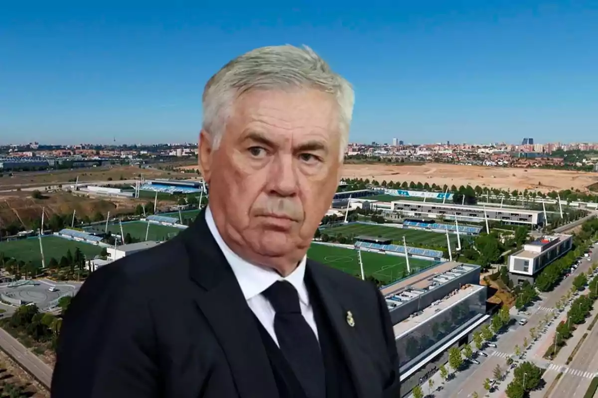 Un hombre de traje frente a un complejo deportivo con campos de fútbol y edificios en un día soleado.