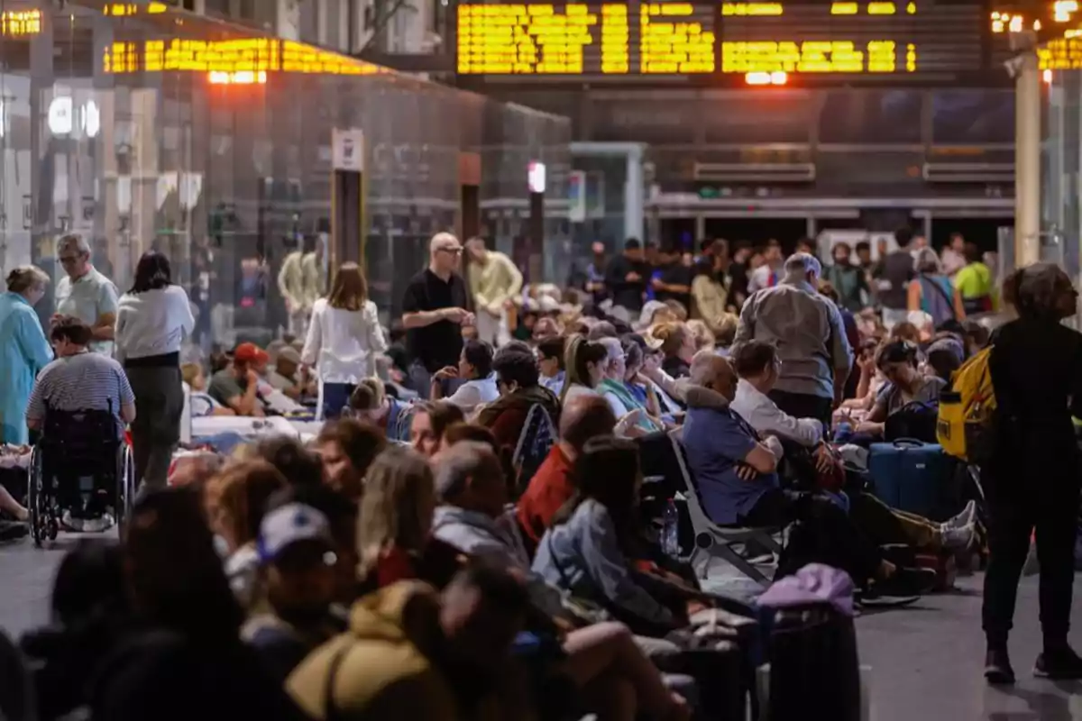 Un grupo numeroso de personas espera en una estación de tren, algunas sentadas y otras de pie, con un tablero de información iluminado al fondo. Un grupo numeroso de personas espera en una estación de tren, algunas sentadas y otras de pie, con un tablero de información iluminado al fondo.