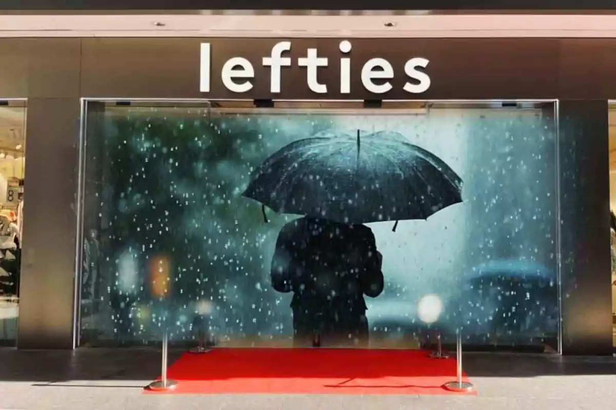 Entrada de una tienda Lefties con un cartel de una persona bajo la lluvia sosteniendo un paraguas y una alfombra roja en el suelo.