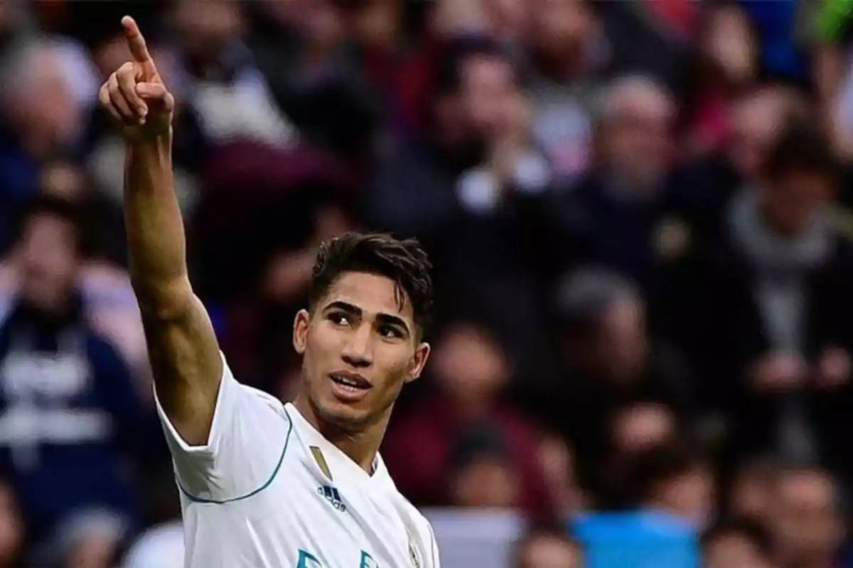 Un jugador de fútbol con uniforme blanco señala con el dedo hacia arriba mientras está en el campo de juego, con una multitud desenfocada en el fondo.