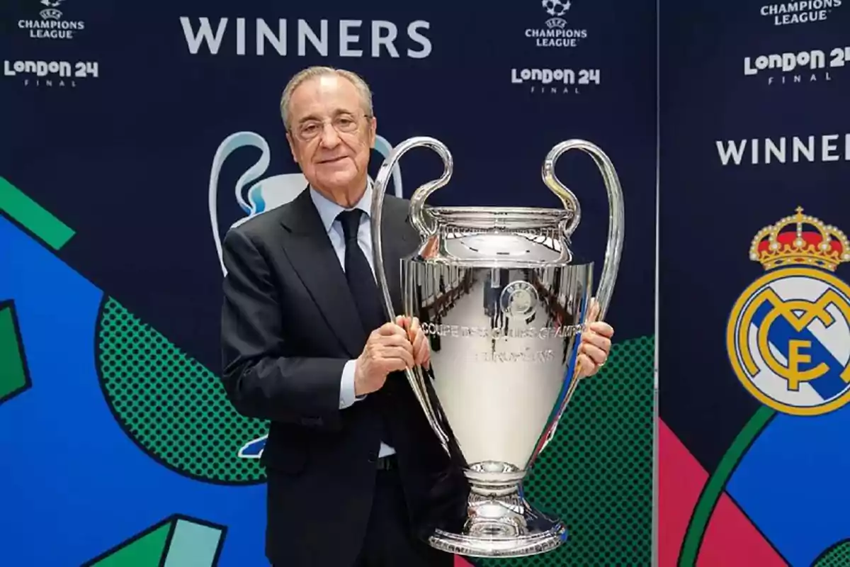 Florentino Pérez sosteniendo el trofeo de la Liga de Campeones de la UEFA frente a un fondo con el logo del Real Madrid y la palabra "WINNERS".