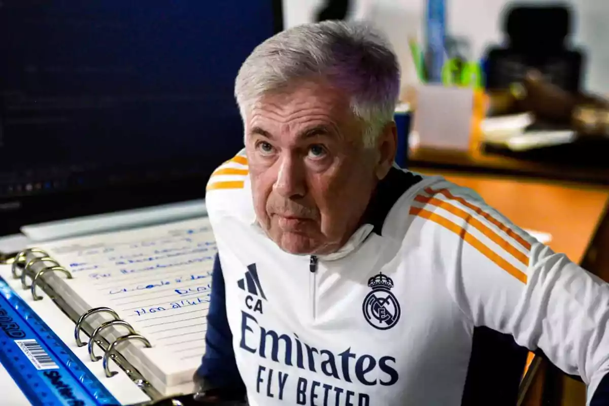 Un hombre con uniforme deportivo del Real Madrid está frente a un escritorio con una computadora portátil y un cuaderno.