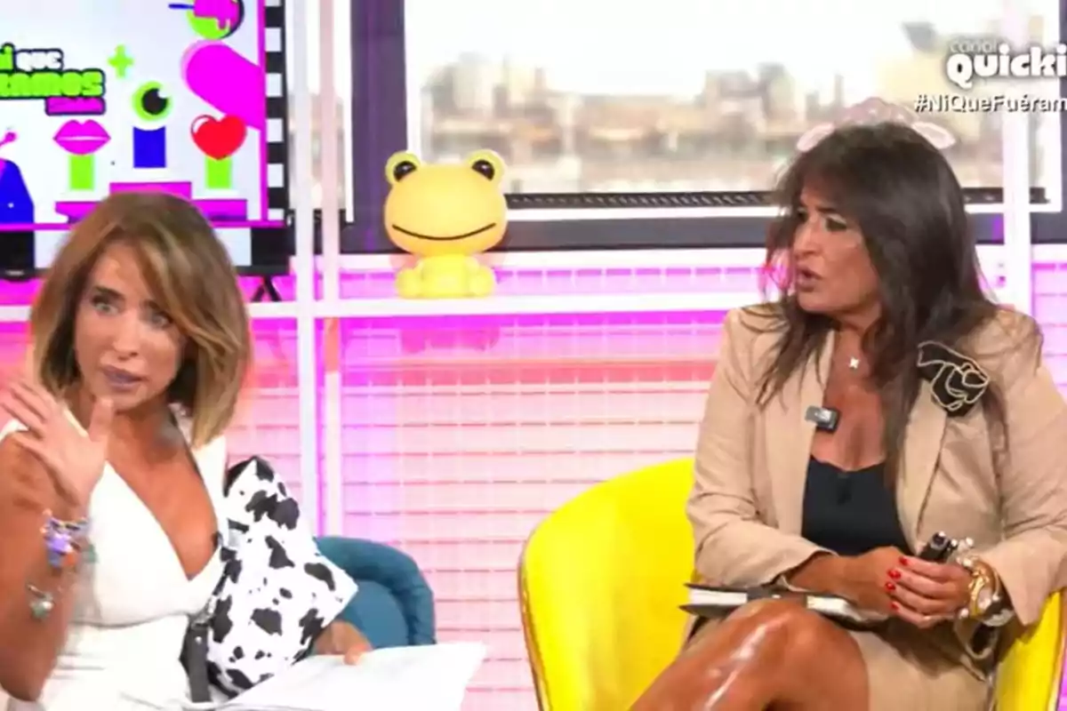 Dos mujeres conversan en un set de televisión colorido con un muñeco amarillo de rana en el fondo.