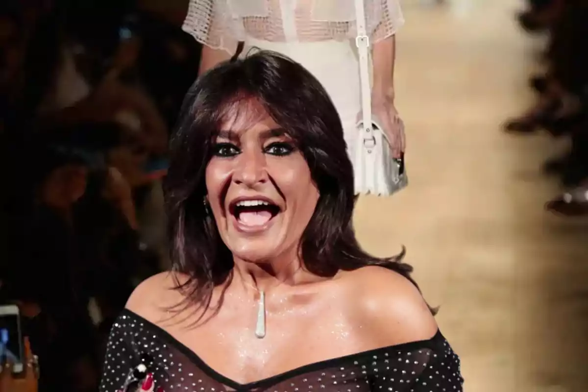 Una mujer sonriente con un vestido negro de hombros descubiertos y fondo de pasarela de moda.