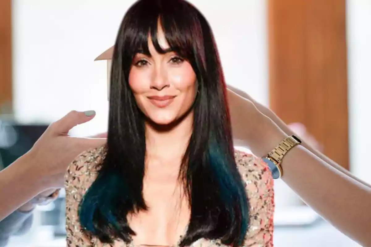 Mujer con cabello largo y lacio de color negro con puntas azules y flequillo, sonriendo mientras alguien sostiene un peine cerca de su cabeza en un entorno de salón de belleza.
