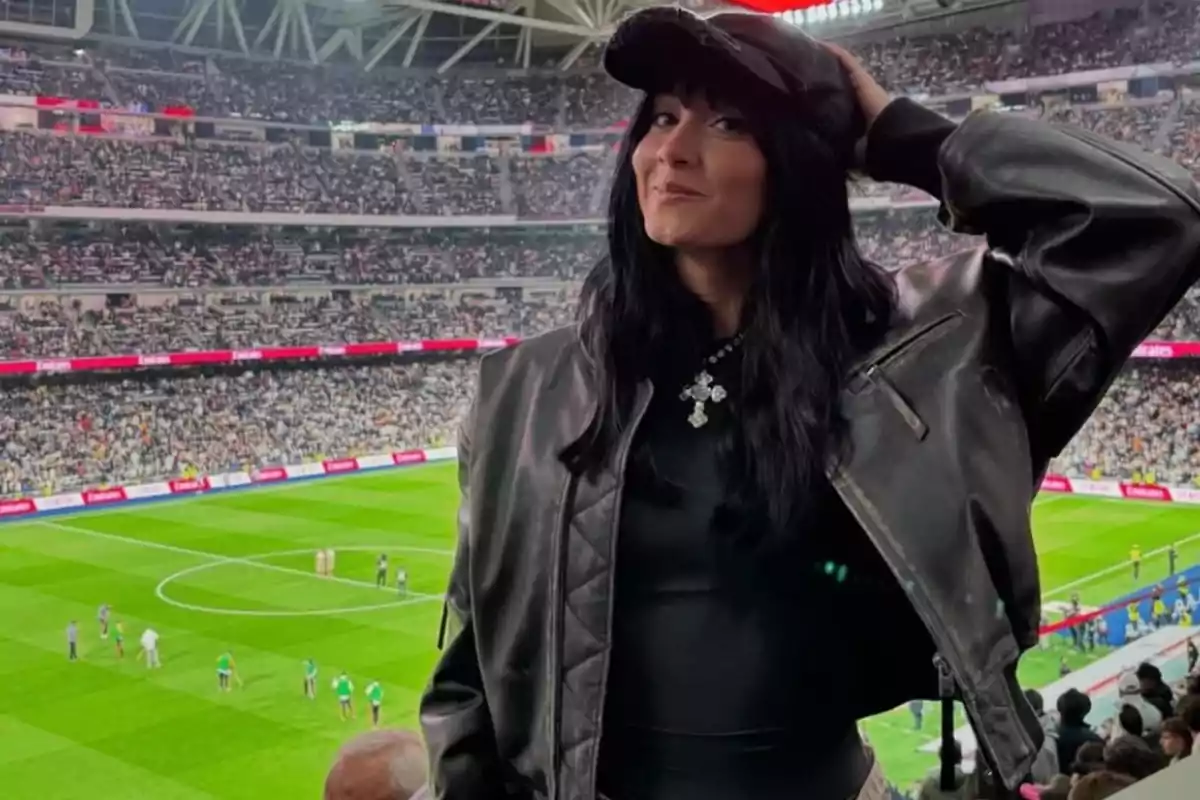 Una persona con chaqueta de cuero y gorra negra posa en un estadio lleno de espectadores con el campo de fútbol al fondo.