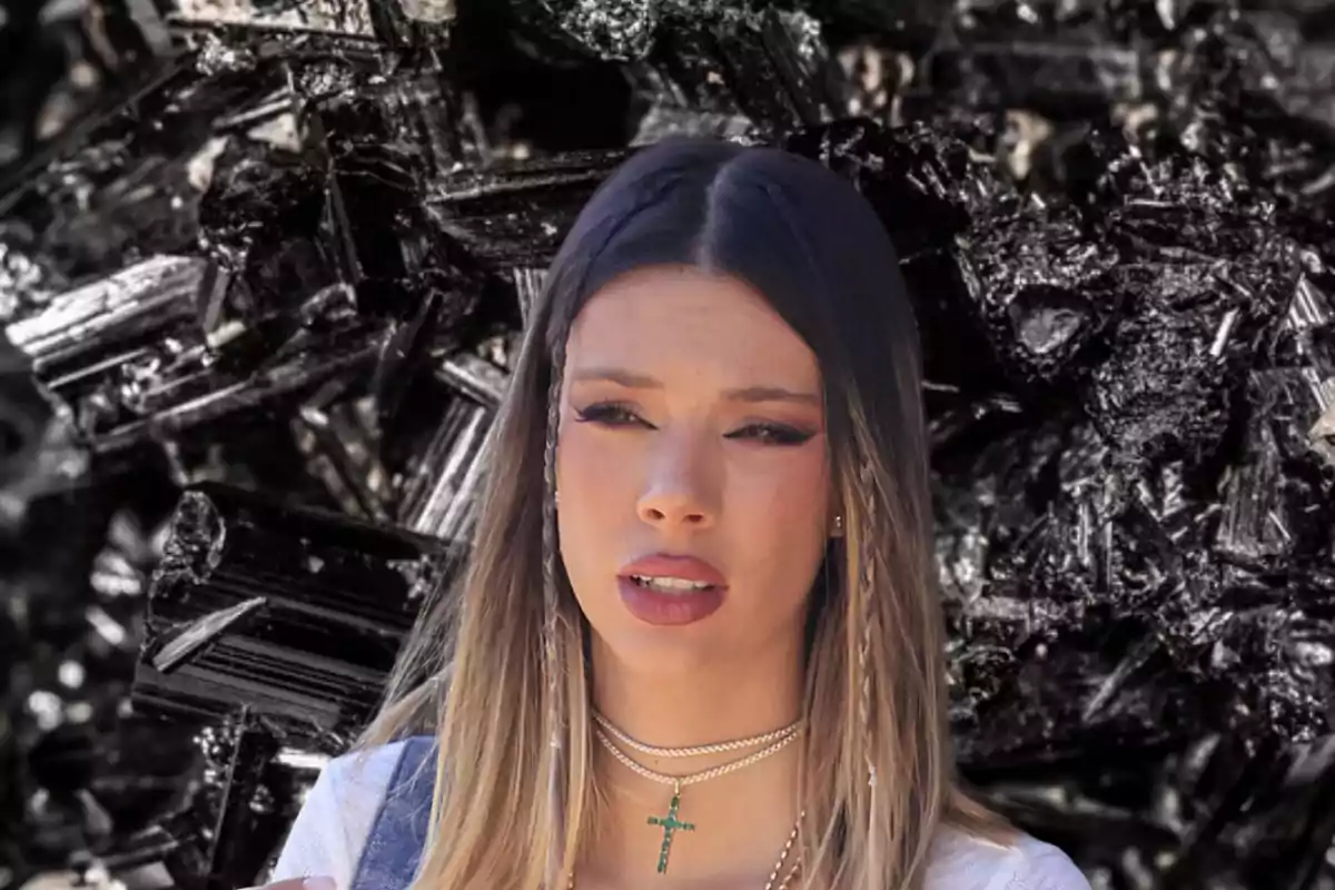 Una mujer con cabello largo y rubio, usando un collar con una cruz verde, está frente a un fondo de cristales negros.