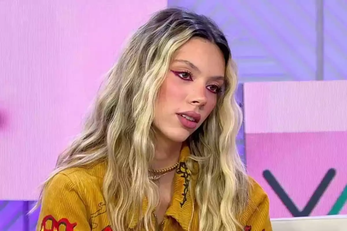Una persona con cabello largo y ondulado, usando una chaqueta amarilla, está en un entorno con fondo rosa y morado.