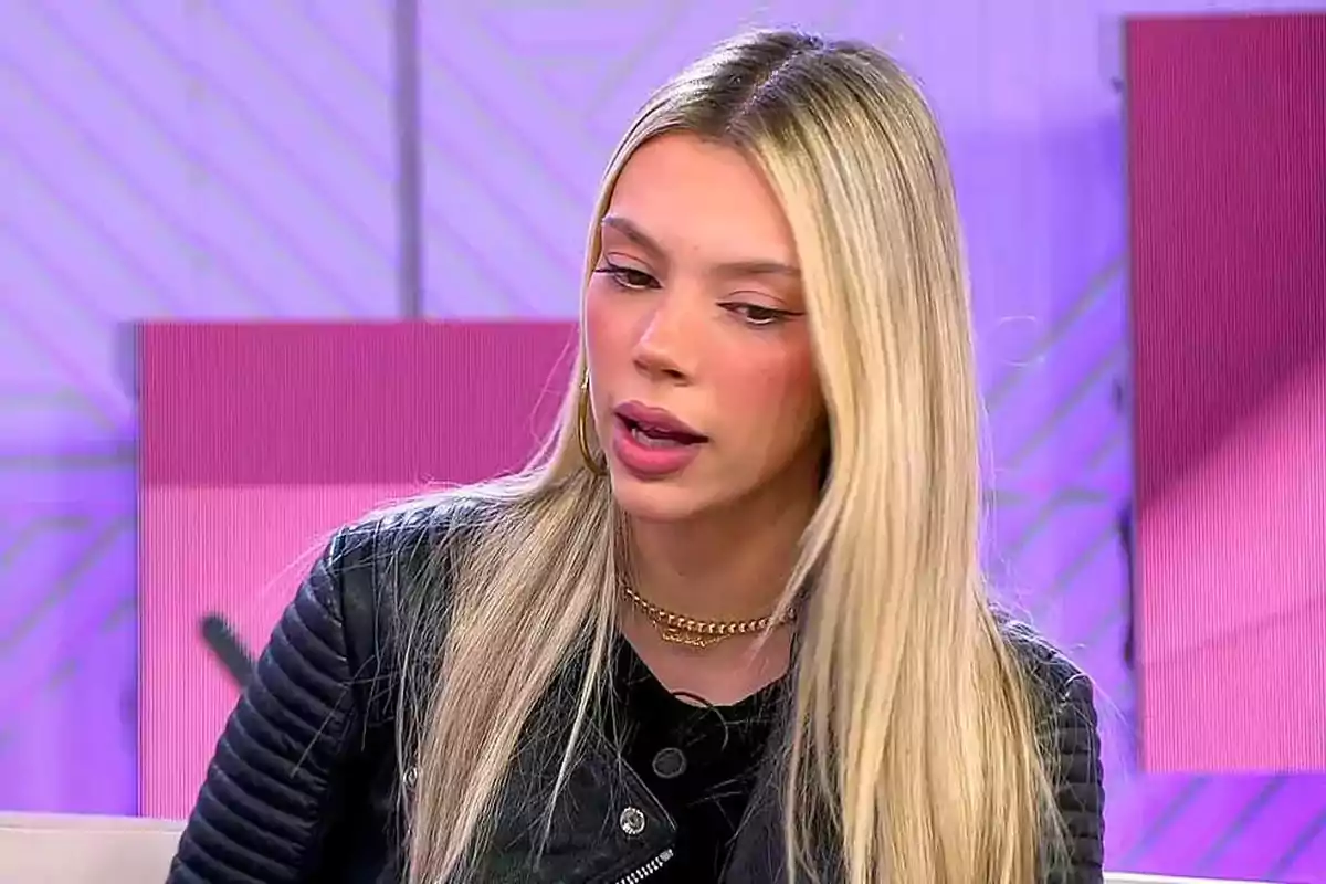 Una persona con cabello rubio y chaqueta negra en un set de televisión con fondo morado. Una persona con cabello rubio y chaqueta negra en un set de televisión con fondo morado.