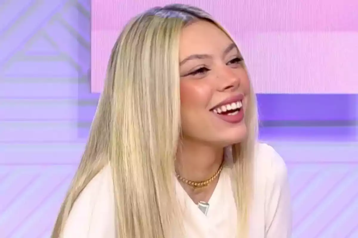 Una persona con cabello rubio sonríe mientras está sentada en un entorno con tonos morados y rosados. Una persona con cabello rubio sonríe mientras está sentada en un entorno con tonos morados y rosados.
