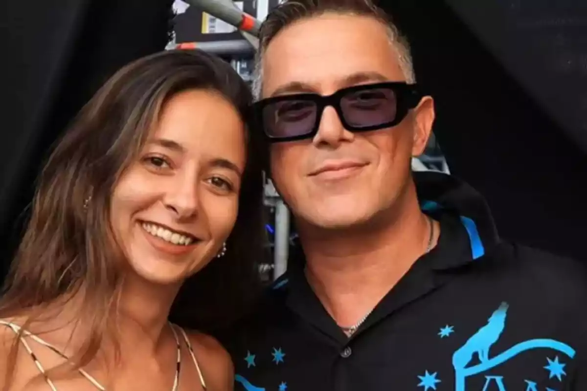 Una mujer joven sonriente junto a un hombre con gafas oscuras y camisa negra con detalles azules posan juntos frente a un fondo oscuro Una mujer joven sonriente junto a un hombre con gafas oscuras y camisa negra con detalles azules posan juntos frente a un fondo oscuro
