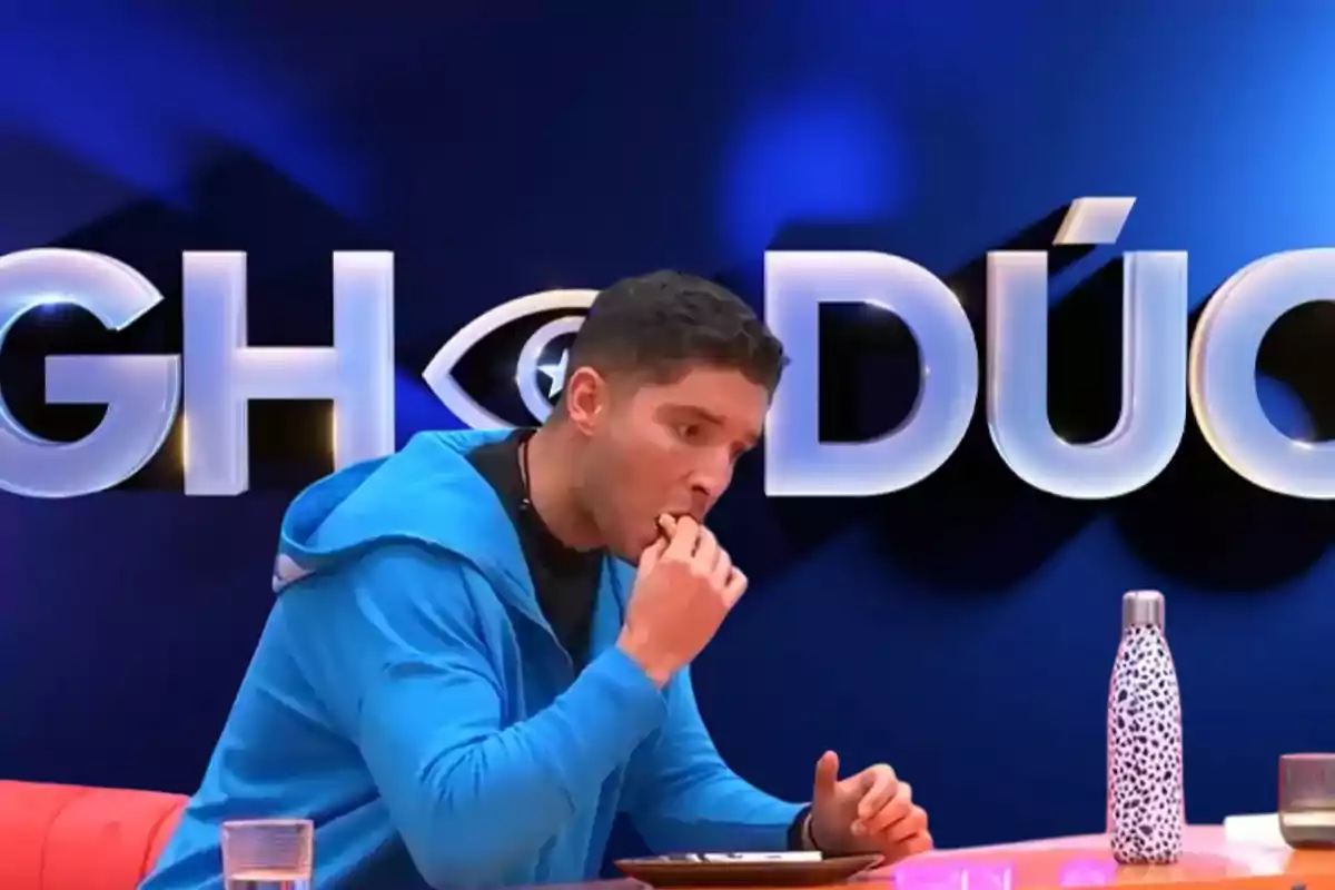 Un hombre con sudadera azul está comiendo frente a un fondo con letras grandes y una botella sobre la mesa.