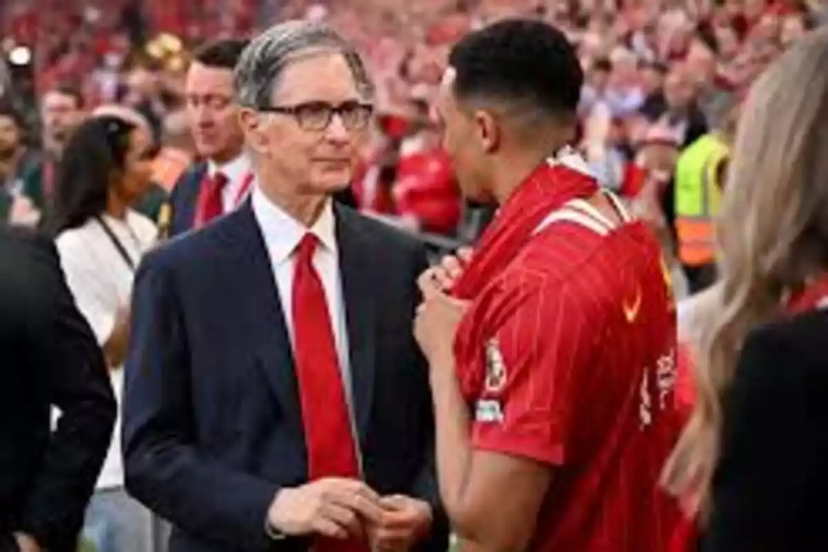 Un hombre de traje y corbata roja conversa con un jugador de fútbol que lleva una camiseta roja en un estadio lleno de espectadores. Un hombre de traje y corbata roja conversa con un jugador de fútbol que lleva una camiseta roja en un estadio lleno de espectadores.