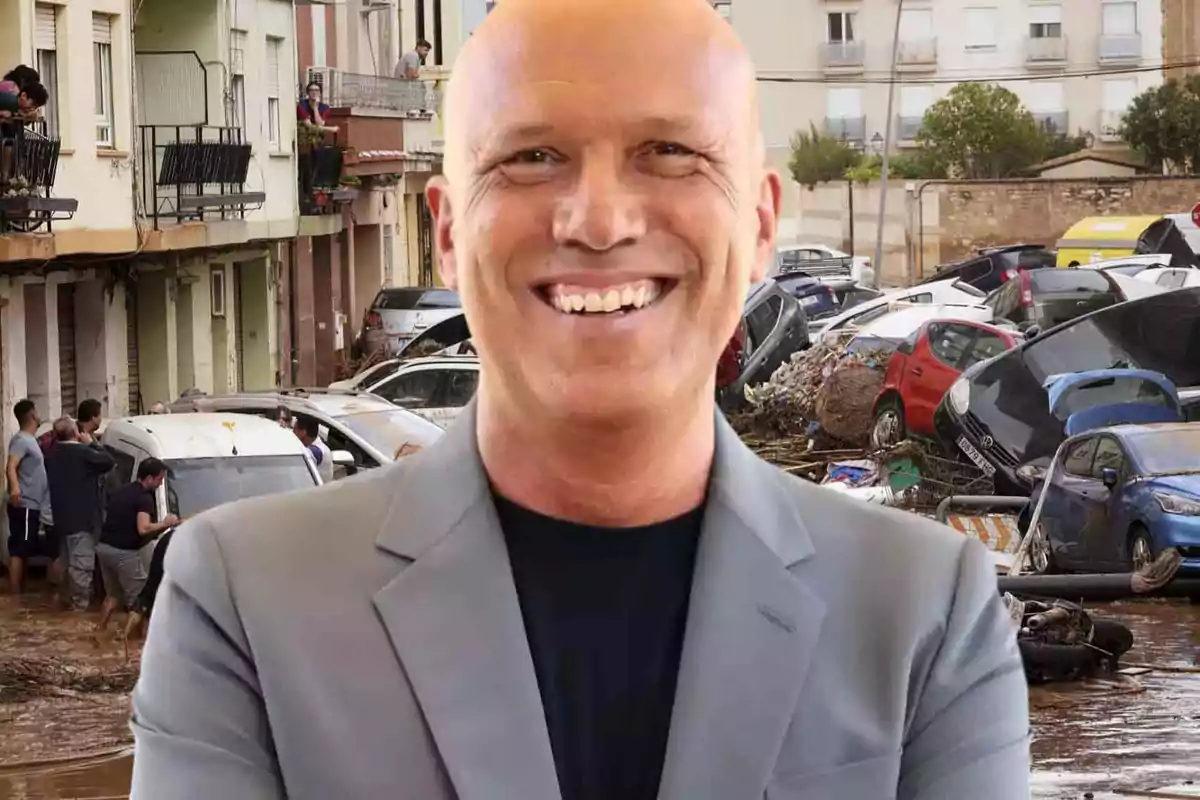 Un hombre calvo sonriente con un traje gris está en primer plano, mientras que en el fondo se observa una calle inundada con coches dañados y personas observando desde los balcones.