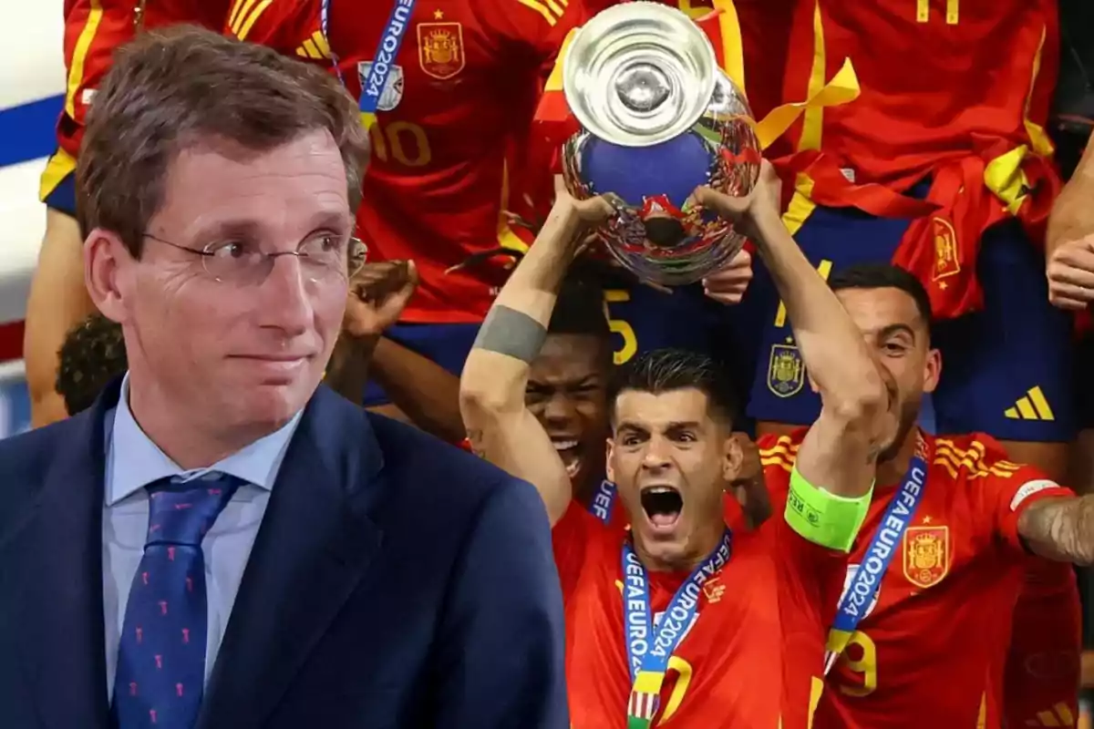 Un hombre con gafas y traje azul está en primer plano, mientras que en el fondo se ve a jugadores de fútbol con uniformes rojos celebrando con un trofeo.